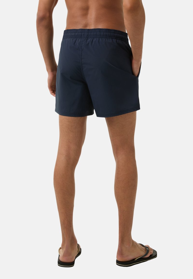 Marineblaue Bad Shorts mit elastischem Bund, seitlichen Taschen und einem glatten, leichten Stoff. Tragen mit schwarzen Flip-Flops.