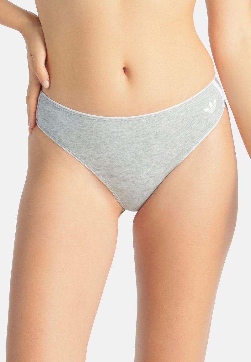 Donna che indossa biancheria intima stile bikini grigio chiaro con un piccolo logo bianco sull'anca sinistra, su uno sfondo chiaro uniforme.