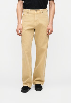 Muž oblečený v béžových nohaviciach s rovnými nohami a čiernych topánkach, stojaci pred jednoduchým svetlým pozadím.