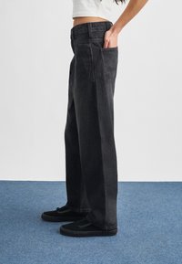 Άτομο που φοράει μαύρο παντελόνι με φαρδιά μπατζάκια, με το χέρι στην πίσω τσέπη, συνδυασμένο με μαύρα αθλητικά παπούτσια και cropped λευκό τοπ.