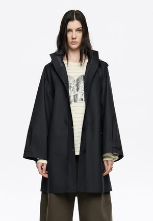 Chubasquero negro impermeable con un corte relajado, que cuenta con un alto collar, grandes botones frontales y dos bolsillos laterales, superpuesto sobre una camiseta de rayas.