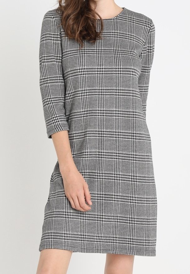Femme portant une robe à carreaux grise et noire longueur genou avec des manches trois-quarts, debout sur un fond blanc uni.