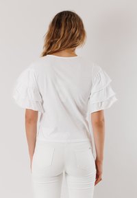 Top bianco a maniche corte con maniche a strati e arricciate. Il retro presenta un semplice scollo rotondo con una texture di cotone liscia.