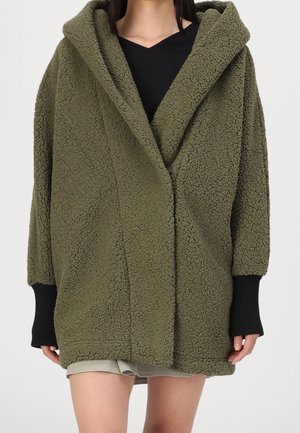 Persoon draagt een oversized olijfgroene sherpa fleece jas met capuchon en zwarte geribbelde manchetten over een zwarte top en beige korte broek.