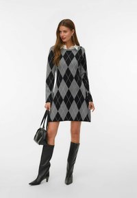 Donna che indossa un vestito a maglione grigio e nero con motivo a rombi, abbinato a stivali neri fino al ginocchio, mentre tiene in mano una borsa nera.