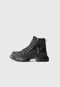 Zwarte enkelboots met een glanzende afwerking, rubberen zool, veter-sluiting en een rits aan de zijkant. Voorzien van een merklabel en textuurdetails langs de zool.