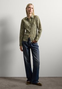 Camicia button-up verde chiaro a fantasia con maniche lunghe, abbinata a jeans scuri di taglio largo e ballerine verde oliva.