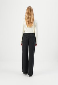 Woman with long blonde hair wearing a white long-sleeve top and high-waisted black wide-leg jeans, standing facing a white wall.