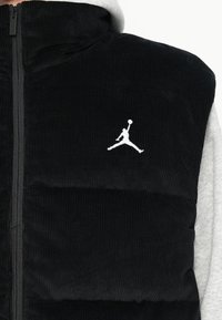 Čierny kordurový vest s horizontálnym prešívaním, obsahuje predný zips a biely Jumpman logo na ľavej hrudi. Šedá mikina pod ním.