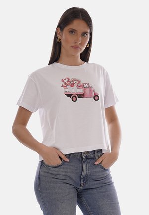 Giovane donna con T-shirt bianca con camion vintage rosa che trasporta fiori a forma di cuore, abbinata a jeans blu, mani nelle tasche.