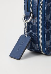 Borsa in denim blu con zip argentata, caratterizzata da una texture a motivo e un'etichetta in pelle blu attaccata da una catena argentata.