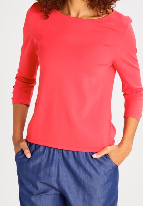 Femme portant un haut corail à manches trois-quarts avec des détails de boutons sur les manches et un pantalon bleu ample, mains dans les poches.