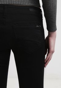 Czarne dżinsy o slim fit, z dwiema tylnymi kieszeniami, subtelnymi przeszyciami i skórzaną naszywką w pasie. Tekstura jest gładka.