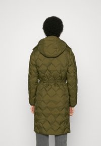 Persona vistiendo un abrigo acolchado largo de color verde oliva con capucha, cintura ceñida, de pie de espaldas contra un fondo blanco.