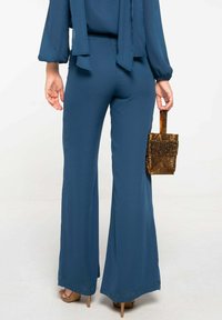 Pantalón acampanado en tejido verde oscuro, con una textura suave. Se sostiene en la mano derecha un pequeño bolso de mano con lentejuelas doradas, que acentúa el conjunto.