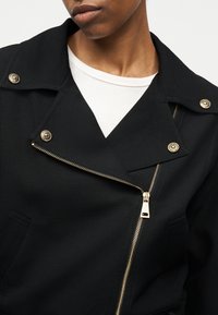 Μαύρο μπουφάν με χρυστό φερμουάρ και κουμπιά φορεμένο πάνω από λευκό τοπ με στρογγυλή λαιμόκοψη, που δείχνει τον άνω κορμό και τον λαιμό ενός ατόμου.