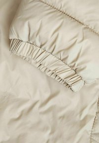 Beige pufferjakke med en blød, skinnende tekstur. Den har en indsamlet elastisk manchet og et glat, slankt stof med subtile syninger.