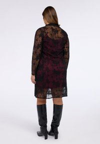 Abito in pizzo nero con motivo floreale, maniche lunghe e sovrapposizione trasparente, con sottoveste bordeaux. Indossato con stivali neri al ginocchio.