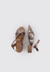 Sandalias de cuero bronce con tiras frontales cruzadas, correa ajustable en el tobillo y un tacón de bloque de madera marrón. Textura suave, diseño casual.
