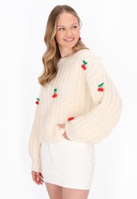 Krémový pletený sveter s oversized rukávmi, zdobený aplikáciami červených a zelených čerešní. Nosený s bielou mini sukňou. Textúrovaná látka.
