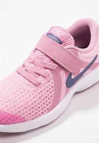 Růžové tenisky Nike Revolution 4 s síťovinovým svrškem, suchým zipem, tkaničkami a námořnickým modrým swoosh na bílé podrážce.