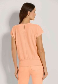 Frau mit langen braunen Haaren trägt ein strukturiertes, pfirsichfarbenes Kurzarmoberteil und passende Hose, steht vor einem hellen Hintergrund mit dem Rücken zur Kamera.