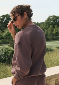 Smeđa majica dugih rukava opuštenog kroja, s rebrastim manšetama i malim logom na leđima. Nošena sa modernim sunčanim naočalama i teksturiranom repom.