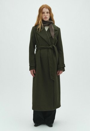 Giovane con capelli rossi che indossa un lungo cappotto verde scuro con cintura, una sciarpa grigia e pantaloni neri su uno sfondo chiaro uniforme.