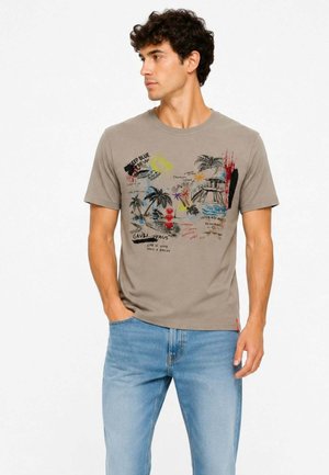 Uomo che indossa una t-shirt a maniche corte color taupe con stampa grafica tropicale e testuale colorata, abbinata a jeans azzurri, guarda verso sinistra.