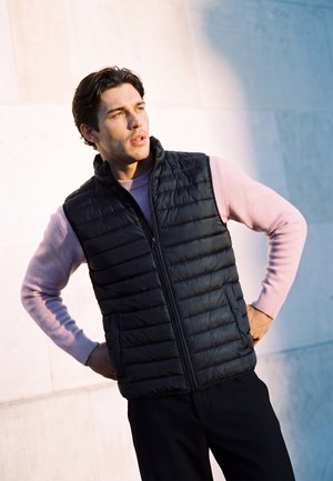 Homme portant une veste matelassée noire par-dessus un pull violet clair, debout les mains sur les hanches, regardant sur le côté avec une expression neutre.