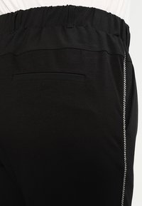 Pantalons noirs avec une taille élastique, présentant un détail de rayure sur le côté. Tissu lisse avec une poche arrière unique. Design simple et fonctionnel.