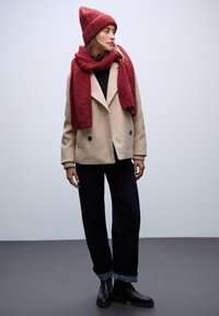 Abrigo beige de doble botonadura, camiseta negra, jeans oscuros con puños doblados, bufanda roja texturizada y gorro a juego. Tejido suave y diseño estructurado.