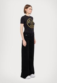 Black cotton t-shirt with gold logo and chain design paired with high-waisted black wide-leg jeans. White sneakers complete the outfit.