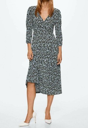 Femme portant une robe midi noire et blanche à motif, manches trois-quarts, avec des chaussures blanches à talons aiguilles, debout.