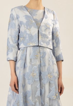Femme portant une robe fleurie bleu clair et une veste courte assortie à manches trois-quarts, avec des accents de feuilles dorées.