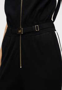 Μαύρη ολόσωμη φόρμα με χρυστό φερμουάρ και υφασμάτινη ζώνη με ορθογώνια αγκράφα. Λευκές πλευρικές ρίγες συμπληρώνουν τον συνολικό σχεδιασμό.