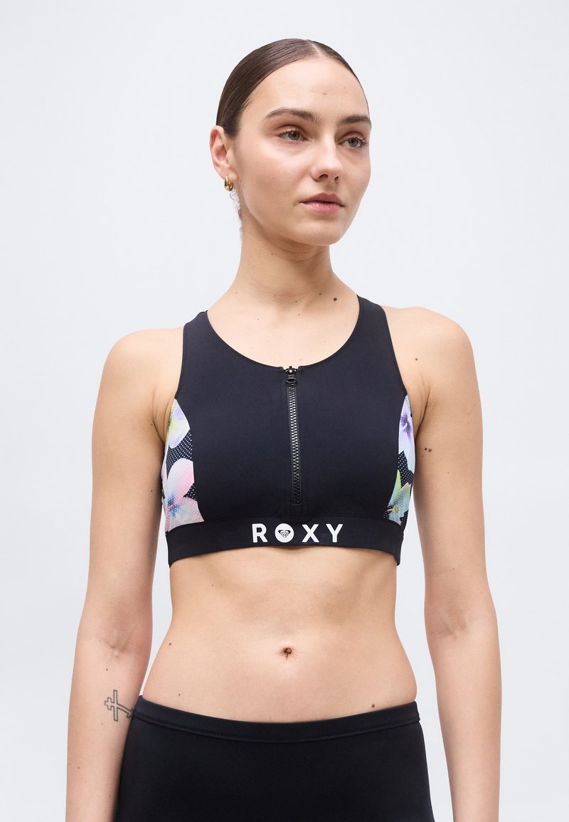 Jeune femme aux cheveux lissés en arrière portant un soutien-gorge de sport noir à fermeture éclair avant avec des panneaux latéraux floraux et le logo "ROXY", debout devant un fond uni.