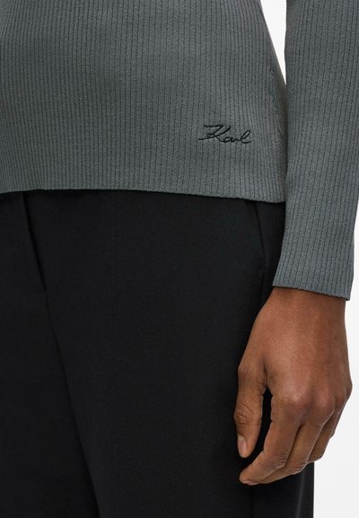 Gros plan d'une main reposant sur le côté, portant un pull gris côtelé avec « Karl » brodé près de l'ourlet, associé à un pantalon noir.