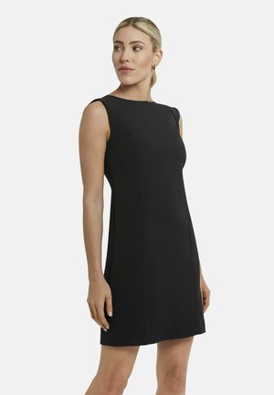 Frau mit blondem Haar, das zu einem niedrigen Pferdeschwanz gebunden ist, trägt ein ärmelloses, schwarzes, knielanges Kleid, steht und blickt nach links.