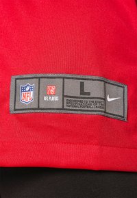 Vörös anyagú, textúrált felülettel, szürke címkével, amelyen szerepel az NFL és a Nike logó, "L" méret, valamint ápolási utasítások.