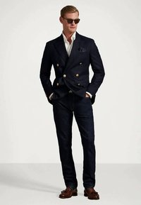 Moški nosi temno dvojno zapončeno suknjič z zlatimi gumbi, belo srajco, temne kavbojke, rjave loaferje, sončna očala in roke v žepih.