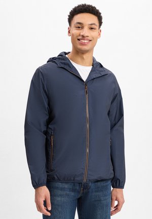Marineblauwe hoodie met een textuurafwerking, voorzien van een rits aan de voorkant, zijzakken en elastische manchetten. Draagt over een wit shirt en jeans.