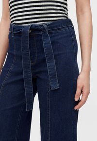 Femme portant un jean taille haute bleu foncé avec une ceinture en tissu nouée devant et un haut rayé noir et blanc.
