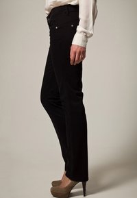 Mustat corduroy-housut, joissa on istuva muotoilu, etutaskut ja vetoketju, yhdistettynä beigellä korkokenkiä ja valkoiseen puseroon.