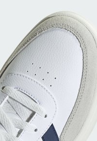 Bijeli kožni tenisice s glatkom teksturom, sivi antilop akcenti, perforirani detalj na prstima i tamnoplava pruga sa strane.