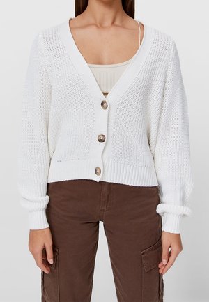 Femme portant un cardigan en maille blanc boutonné sur un débardeur côtelé beige et un pantalon cargo marron foncé.