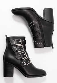 Botas de tobillo de imitación de cuero negro con tacón de bloque, que cuentan con cuatro correas horizontales decoradas con tachuelas y un zipper lateral para el cierre.