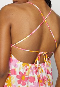 Meca de flores em vestido de verão com padrões florais em rosa e amarelo, alças cruzadas ajustáveis e cintura franzida com um detalhe de laço.