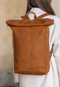 Rudas odinis roll-top kuprinė su lygiu paviršiumi, detalia siuvimo linija ir viršutine rankena. Kompaktiškas dizainas, tinkantis kasdieniam naudojimui.