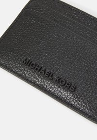 Crni kožni nosač za kartice s teksturiranom površinom, više utora za kartice i utisnutim logotipom "MICHAEL KORS". Kompaktni, pravokutni oblik.